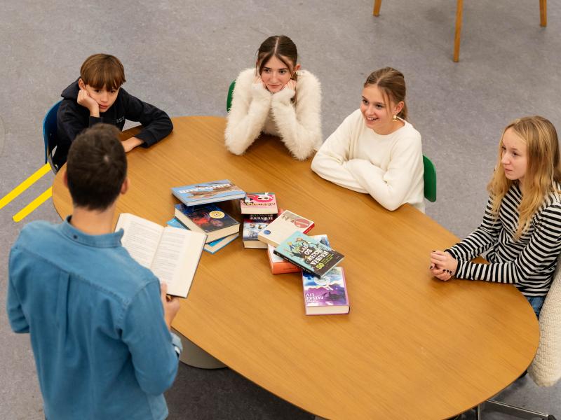 4 unge sidder ved et bord og lytter til en mand der står og læser op af en bog